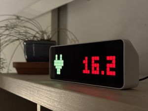Close-up photograph of a white plastic Ulanzi clock, with a black screen made of a 8x32 LED matrix. On the left of the matrix is a shape made of yellow pixels that resembles a power plug and on the right the number 16.2 in red.